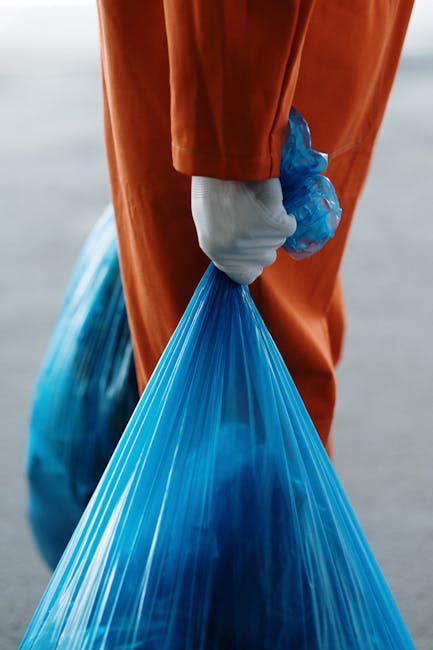 Close-up of a person wearing an orange protective suit and a white glove, holding a tied blue plastic rubbish bag by the top with fingers grasping it firmly. The bag appears to be filled with waste an